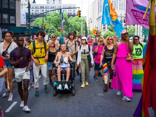 A group including wheelchair users moves down a street with rainbow flags.