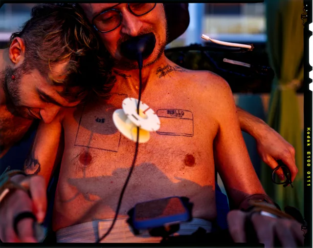 A shirtless wheelchair user with medical tubing reclines under red-orange light.