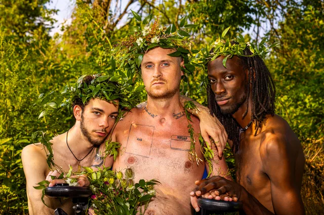Three shirtless people pose closely together outdoors with leafy plants draped across their bodies.