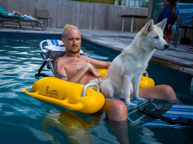 A shirtless wheelchair user sits in a pool beside a white dog and yellow float.