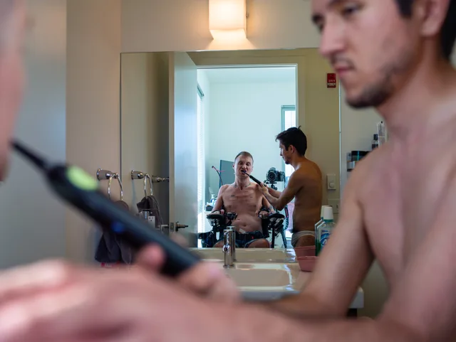 A bathroom mirror reflects a wheelchair user and a nude person standing beside a sink.