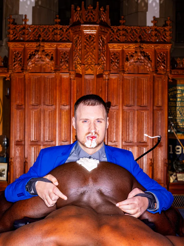 A person in a blue jacket and bow tie sits before carved wood with a nude dark-skinned person lying across their lap.