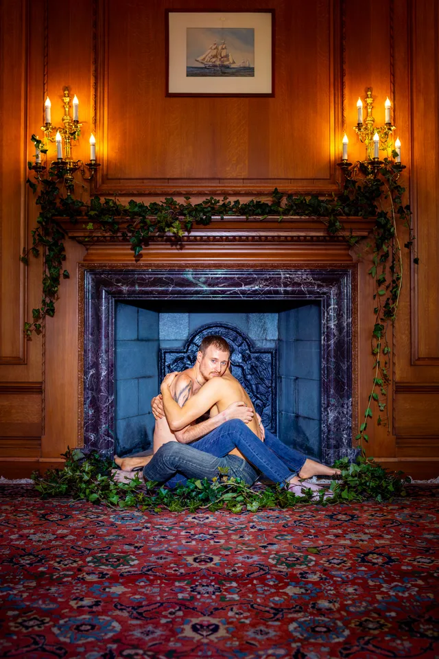 A person sits curled inside an ornate wooden fireplace beneath lit candles.