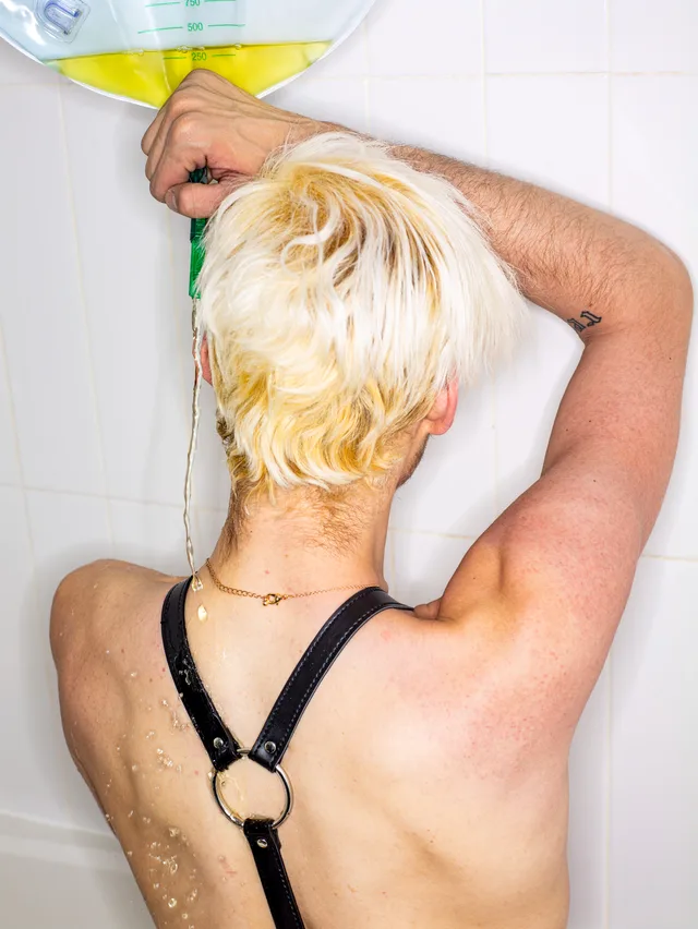 The wet back of a person wearing a black leather harness fills a tiled shower.