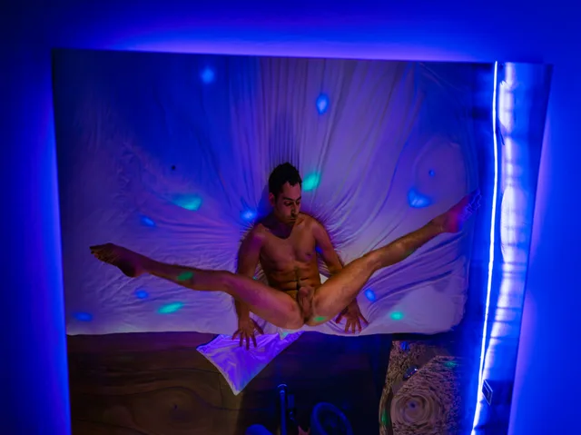 Overhead view of two nude people posed on a white bed under saturated blue and violet light.