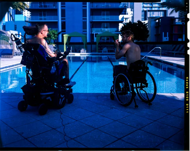 Two wheelchair users face each other beside a pool under deep blue light.