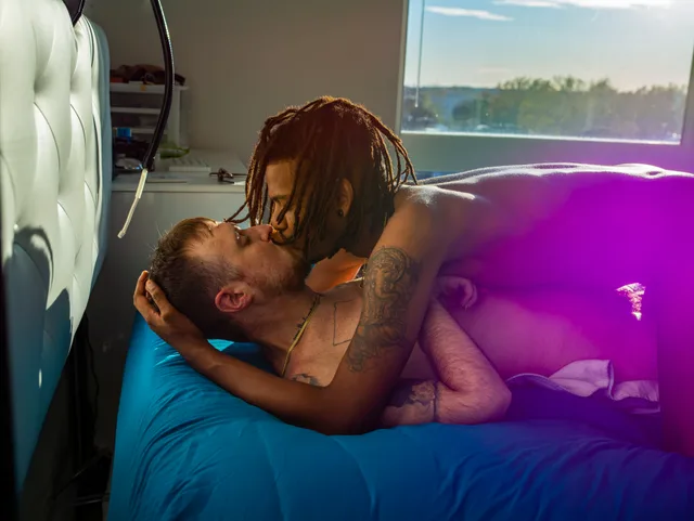 Two nude people lie on a blue bed, one leaning over the other's chest.