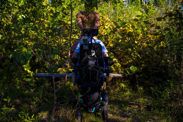 A wheelchair user sits on a narrow path surrounded by dense green foliage.