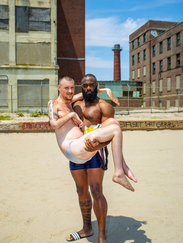 A shirtless dark-skinned person carries a nude pale person across sand near brick buildings.