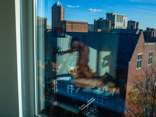 A nude figure is reflected in a window overlooking brick buildings and a city skyline.