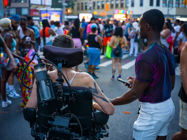 A person walks beside a wheelchair user with camera equipment on a crowded street.
