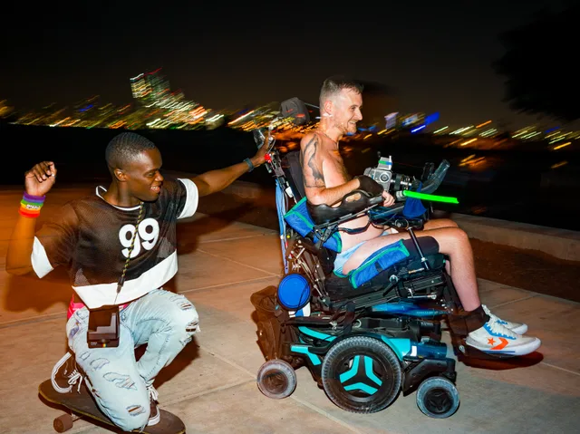 A person crouches while holding hands with a wheelchair user outdoors at night.