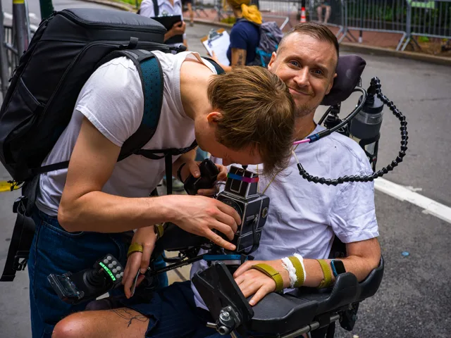 A person bends close to a wheelchair user seated outdoors.
