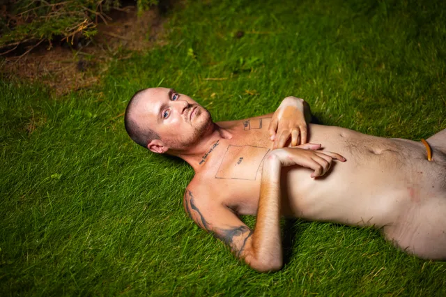 A nude person lies on green grass with hands resting across their chest.