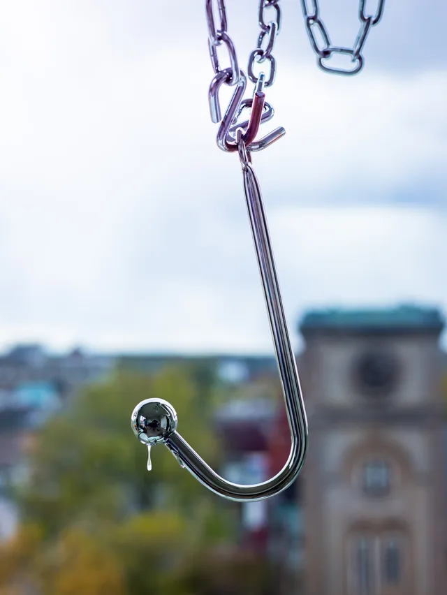A polished metal hook and chains hang before a softly blurred window view, with a droplet suspended from a chrome ball.