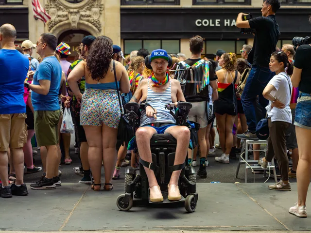 A wheelchair user faces a dense sidewalk crowd beneath a COLE HAAN sign.