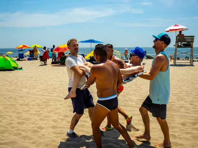 A group gathers around a nude person being carried across a sandy beach.
