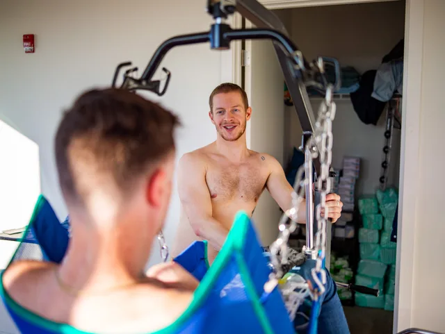 A shirtless person smiles toward a seated person amid ceiling-lift equipment.