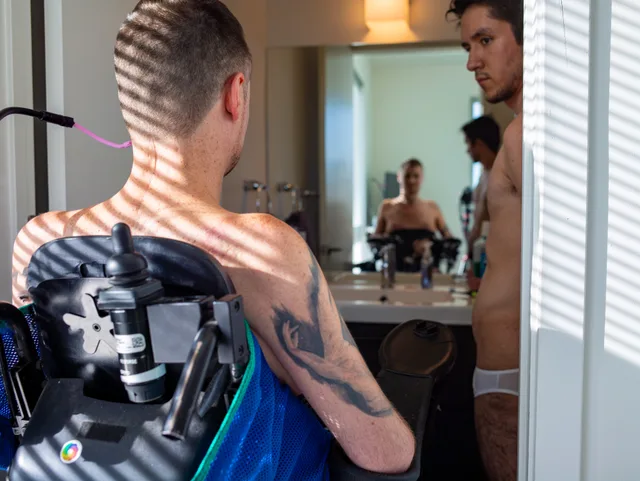 The back of a wheelchair user with a mounted camera faces a mirror reflecting a nude person.