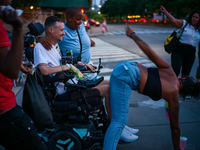 A person lifts one leg beside a wheelchair user while others watch on a city street.