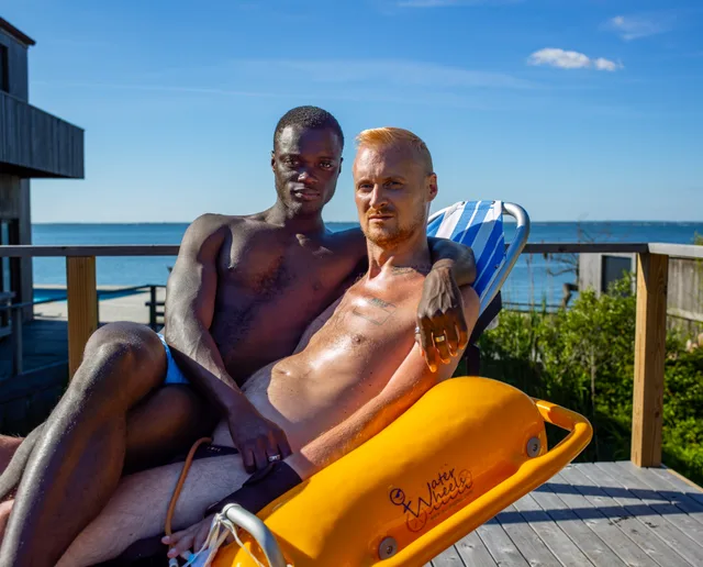 Two nude people sit together on a deck overlooking blue water beside a yellow flotation device.