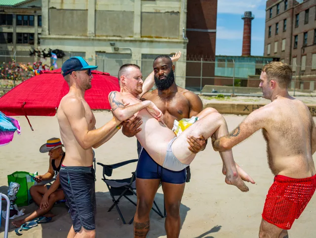 Several shirtless people carry a nude person horizontally on a crowded beach.