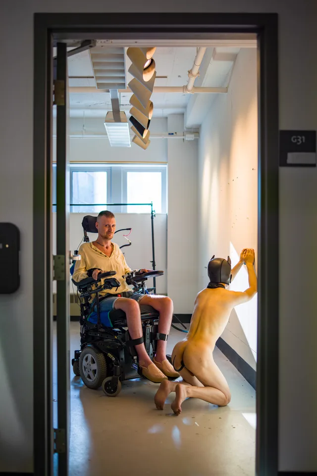 Seen through a doorway, a nude kneeling person raises one arm beside a wheelchair user.
