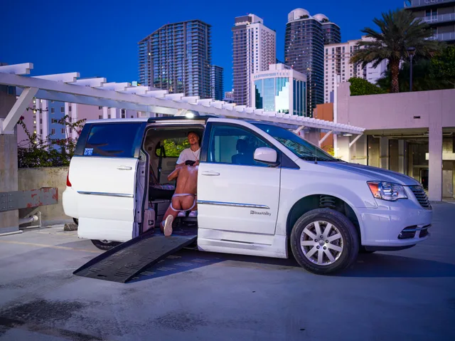 A wheelchair user sits in the open doorway of a white van beneath tall buildings.