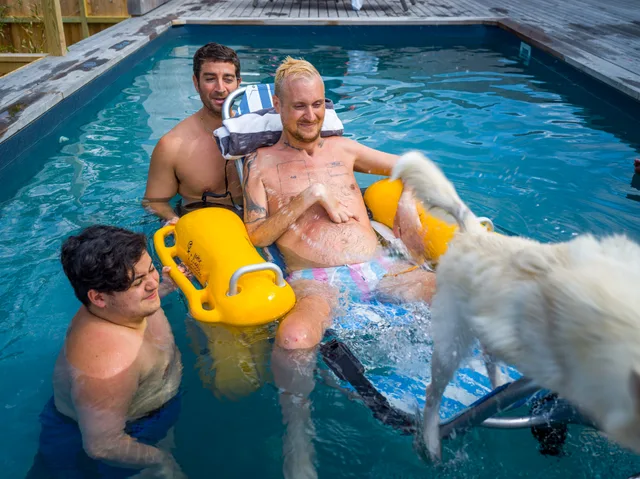 Two people assist a shirtless wheelchair user in a pool beside a white dog and yellow float.