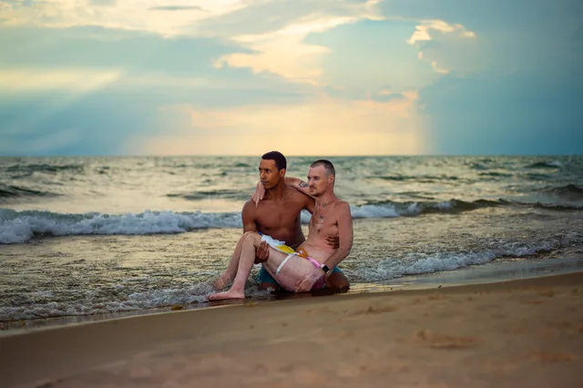 Two nude people sit together at the edge of the ocean beneath a cloudy sky.