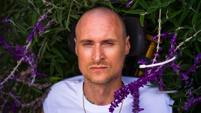 Close portrait of a bald person surrounded by leafy branches and purple flowers.