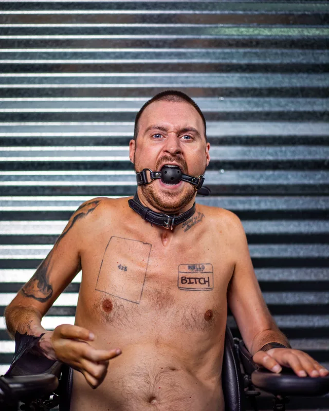 A shirtless wheelchair user wears a black mouth gag and a chest tattoo reading 'BITCH' before a corrugated metal wall.