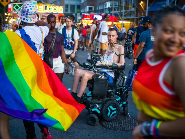A wheelchair user sits within a Pride crowd as a rainbow flag fills the foreground.