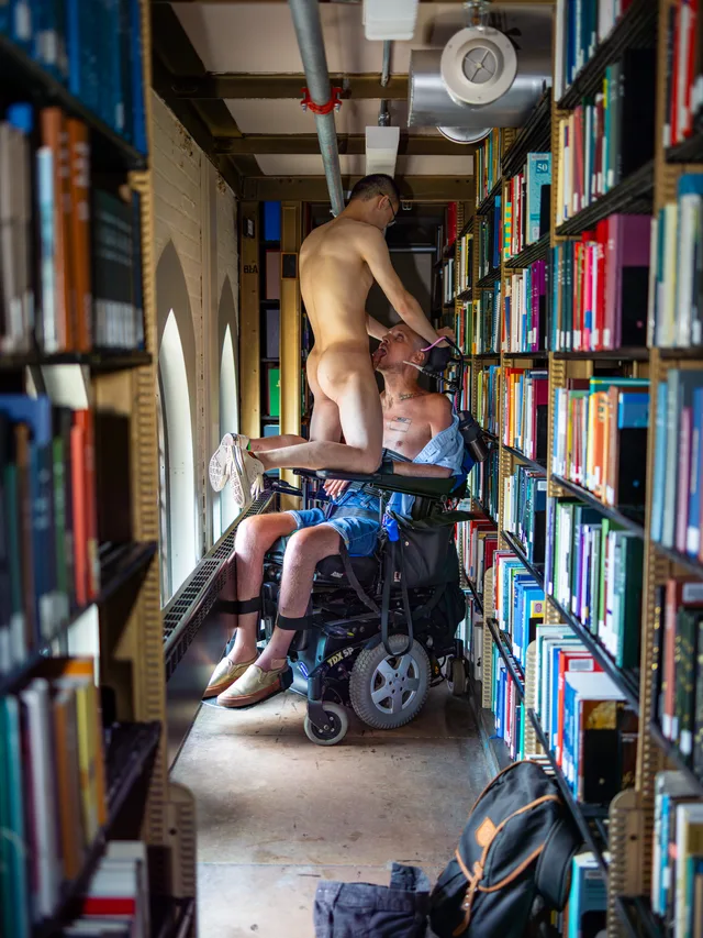 A nude person stands on a library ladder above a wheelchair user in a narrow aisle of books.