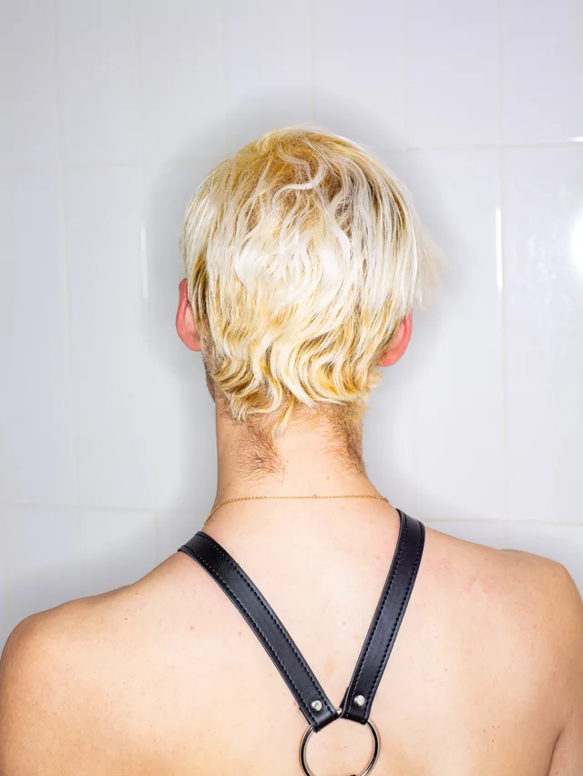 A person with short bleached hair faces a tiled shower wall wearing a black leather harness.