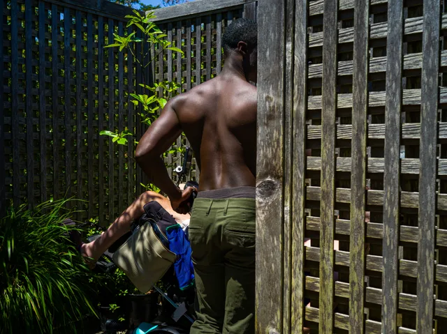 A nude Black person stands with their back to the camera beside an outdoor shower.